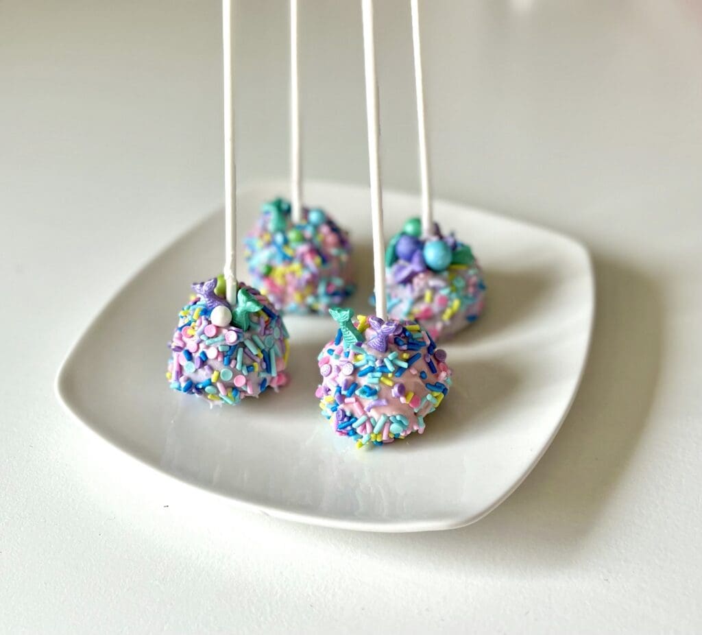 four cake pops with mermaid sprinkles on white plate